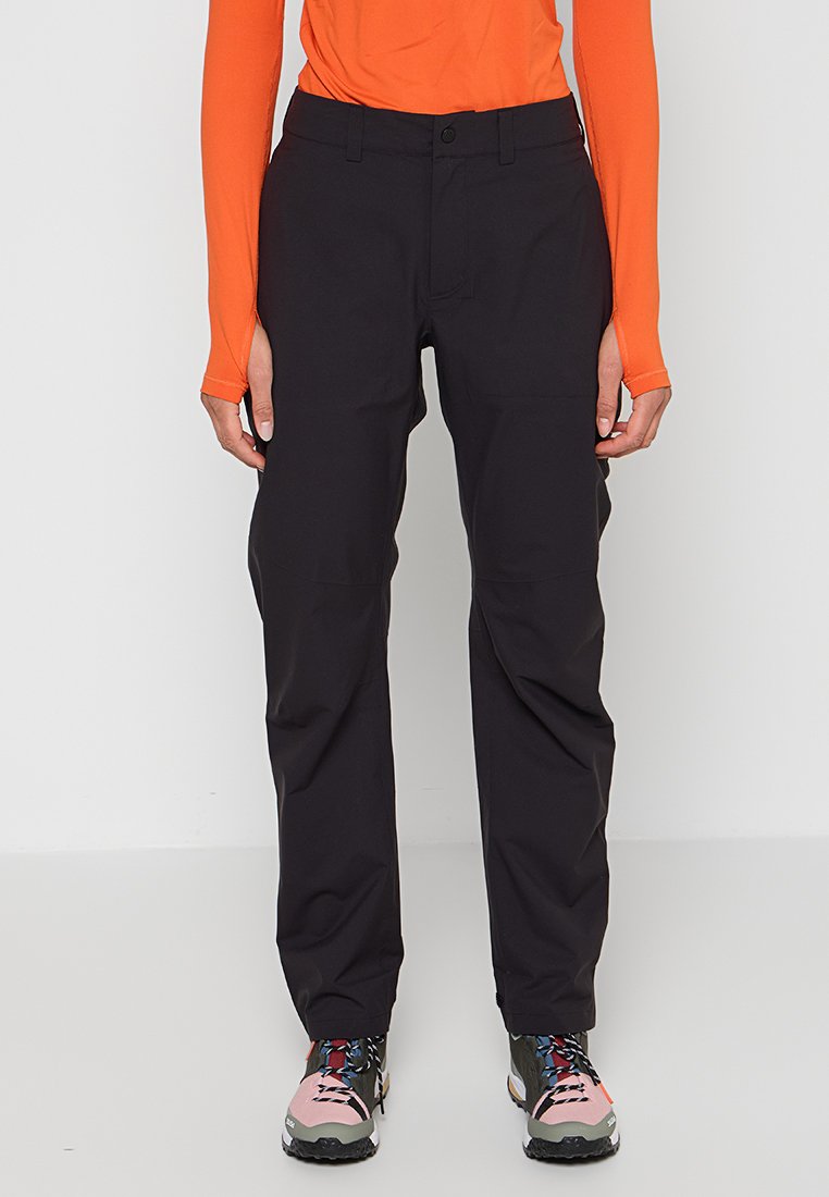 Person trägt schwarze Hosen mit geradem Bein, orangefarbenes Langarmshirt und pink-schwarze Wanderschuhe, steht vor einem einfarbigen Hintergrund.