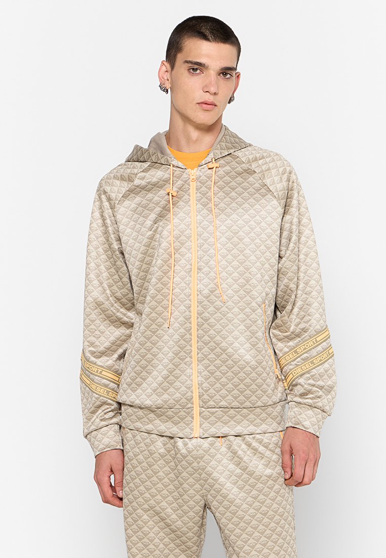 Beige gesteppte Zip-Jacke mit Kapuze, versehen mit orangen Akzenten und gestreiften Marken, kombiniert mit passenden Hosen aus glattem Stoff.