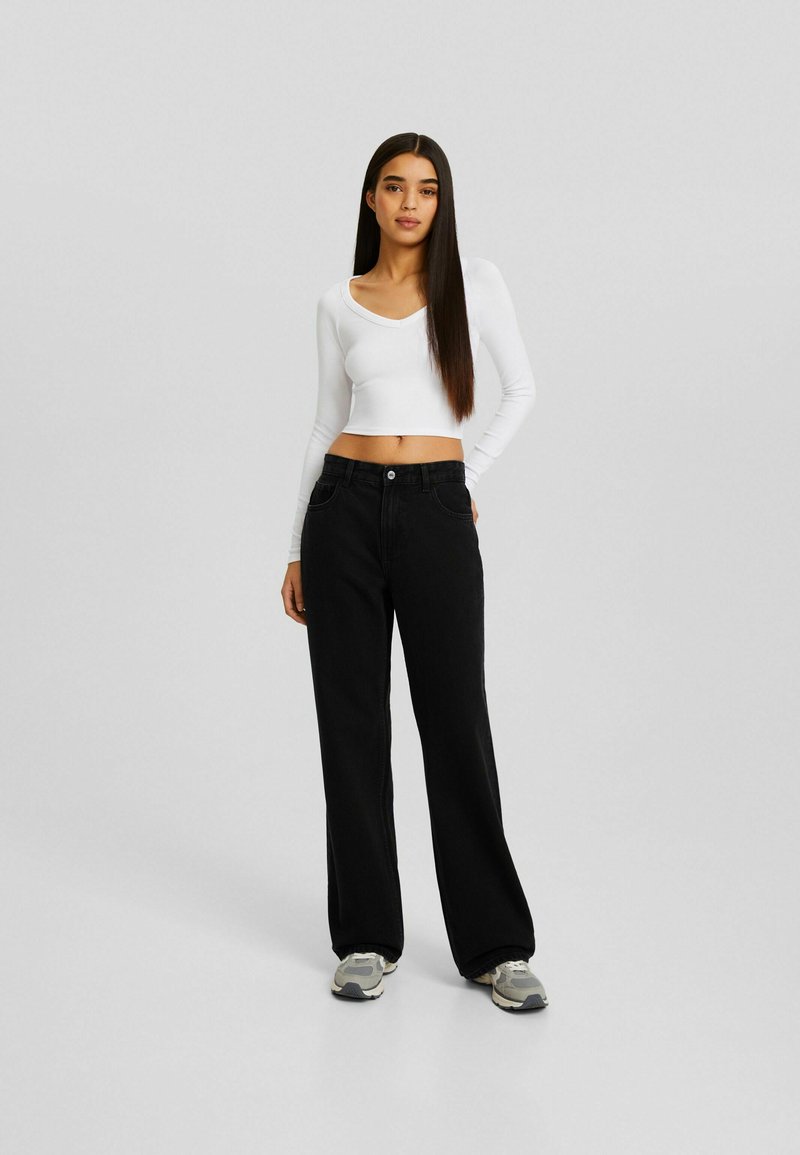 Schwarze weit geschnittene Jeans aus Denim, kombiniert mit einem engen weißen Langarmoberteil. Das Outfit wird mit grauen Sneakers abgerundet.