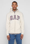 Junger Mann trägt eine cremefarbene Zip-up-Hoodie von GAP über einem karierten Hemd und blauen Jeans, steht vor einem schlichten grauen Hintergrund.