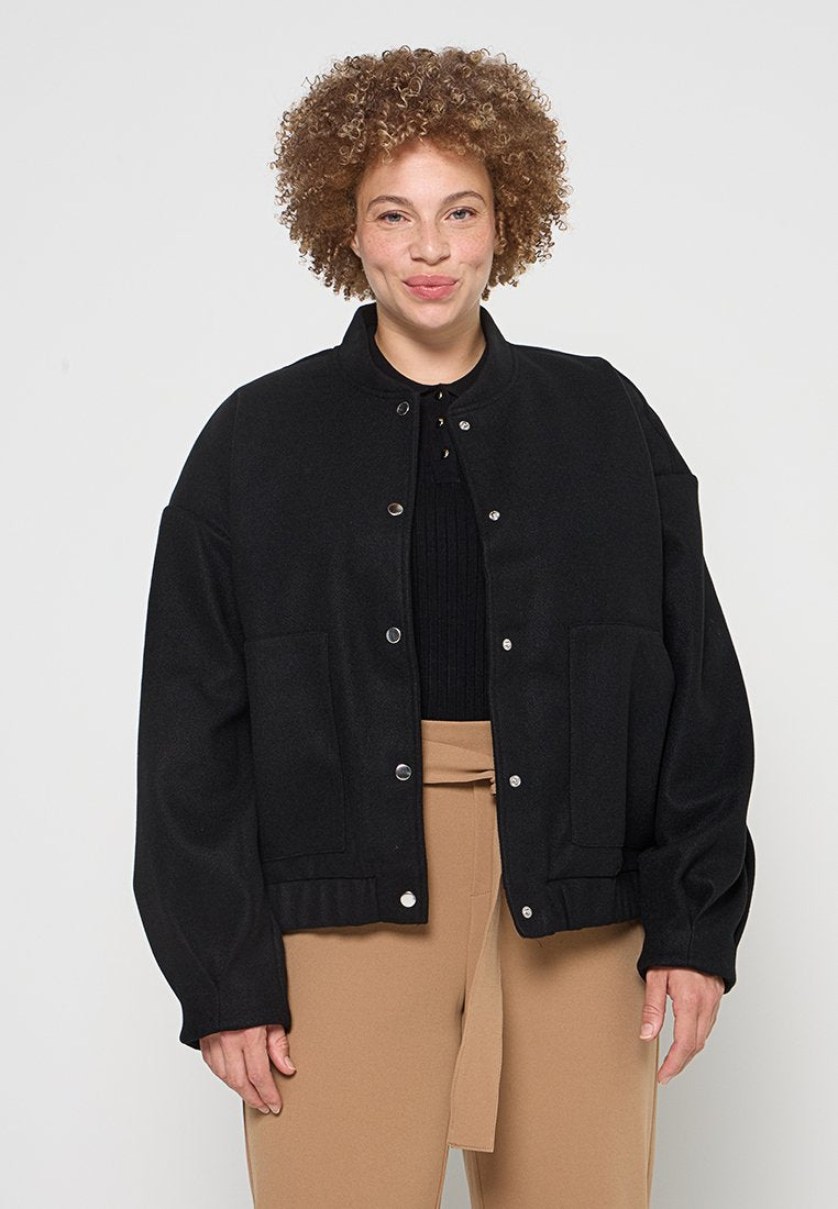 Schwarze Wollmischungjacke mit Rundhalsausschnitt, Druckknöpfen, oversize Passform und seitlichen Taschen, kombiniert mit einem gerippten schwarzen Oberteil und beigen Hosen.