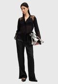 Schwarze transparente Bluse mit Spitzendetails, Knopfverschluss vorne und langen Ärmeln, kombiniert mit schwarzen satinierten weiten Hosen und einer metallisch silbernen Tasche.