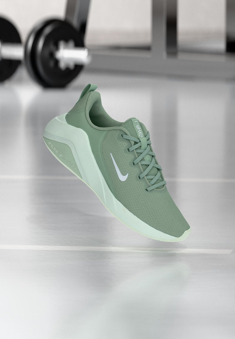 Hellgrüner Nike-Sneaker mit weißem Swoosh-Logo, atmungsaktivem Mesh-Obermaterial und dicker Sohle vor verschwommenem Fitnessgeräte-Hintergrund.