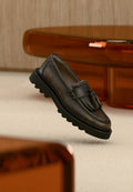 Dunkelbrauner Leder-Slipper mit Quasten, schwarze klobige Sohle, auf beigem Teppich neben durchsichtigem orangefarbenem Möbelstück präsentiert.