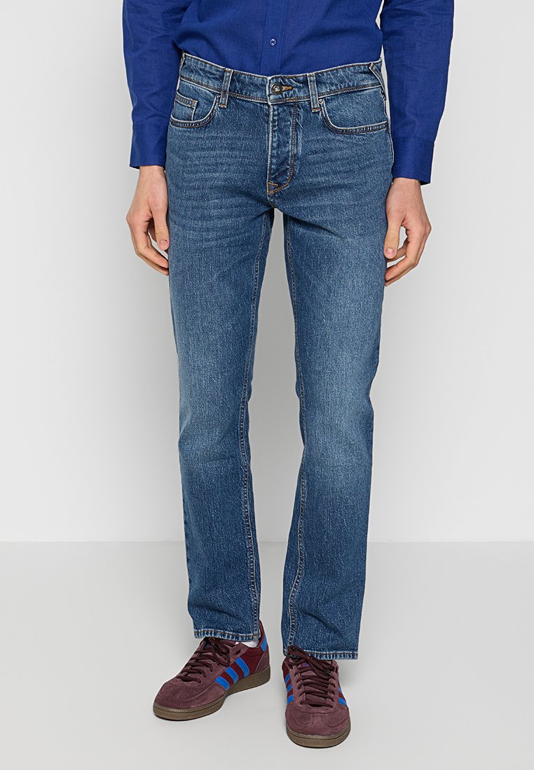 Denim-Jeans in einer mittelblauen Waschung, mit geradem Schnitt, fünf Taschen und dezenten Nähten. Getragen mit burgunderfarbenen Sneakers.