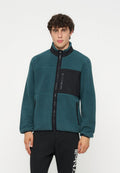 Junger Mann mit lockigem Haar, der eine türkisfarbene Fleecejacke mit schwarzen Reißverschlusstaschen und schwarze Hose mit weißem Logo am linken Bein trägt.