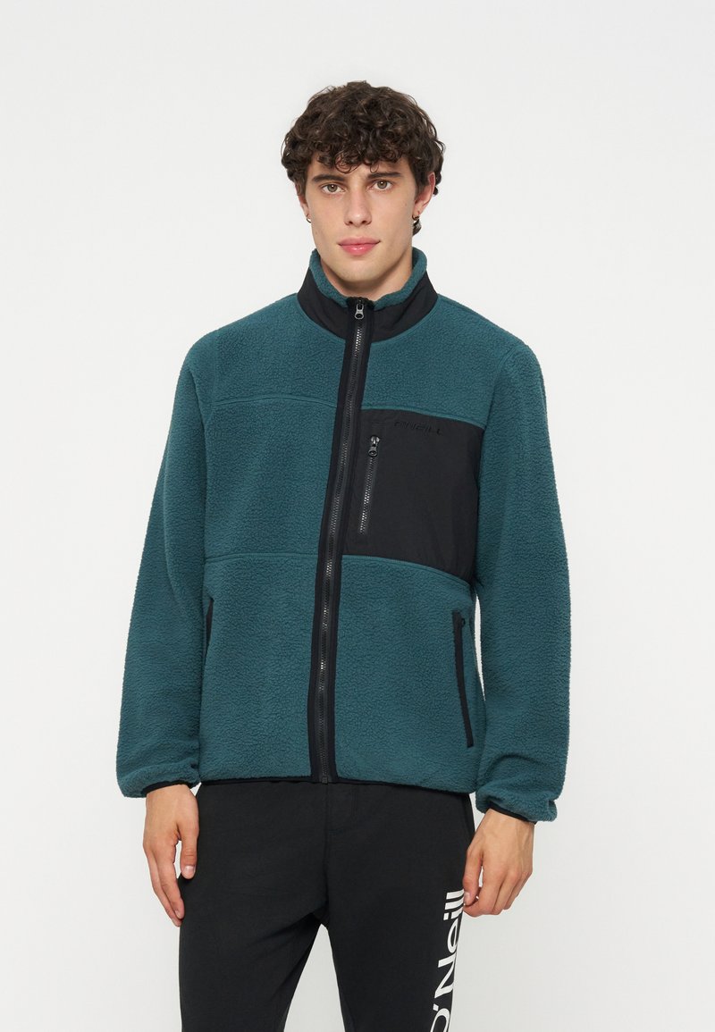Junger Mann mit lockigem Haar, der eine türkisfarbene Fleecejacke mit schwarzen Reißverschlusstaschen und schwarze Hose mit weißem Logo am linken Bein trägt.