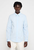 Mann in einem hellblauen langärmeligen Button-Up-Hemd und weißen Hosen, der mit dem Gesicht zur Kamera vor einem schlichten weißen Hintergrund steht.