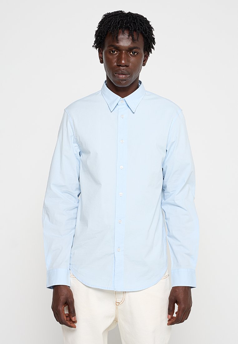 Mann in einem hellblauen langärmeligen Button-Up-Hemd und weißen Hosen, der mit dem Gesicht zur Kamera vor einem schlichten weißen Hintergrund steht.