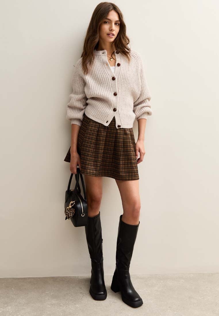 Beige Rippstrick-Cardigan mit braunen Knöpfen, kombiniert mit einem braunen karierten Rock und kniehohen schwarzen Stiefeln mit Absatz, ergänzt durch eine schwarze Handtasche.