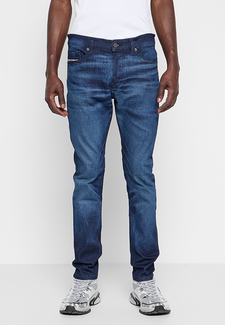 Dunkelblaue Denim-Jeans mit Slim-Fit, ausgestattet mit einem Fünf-Taschen-Design und einem Knopfverschluss, getragen mit grauen Sportschuhen.