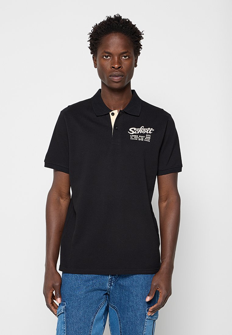 Mann trägt ein schwarzes Polohemd mit weißem Logo-Schriftzug auf der linken Brust und blaue Denim-Cargohosen, steht vor einem einfarbigen Hintergrund.