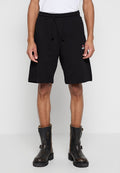 Mann trägt schwarze Shorts mit Kordelzug und schwarze Leder-Stiefeletten, steht vor einem einfarbigen Hintergrund.