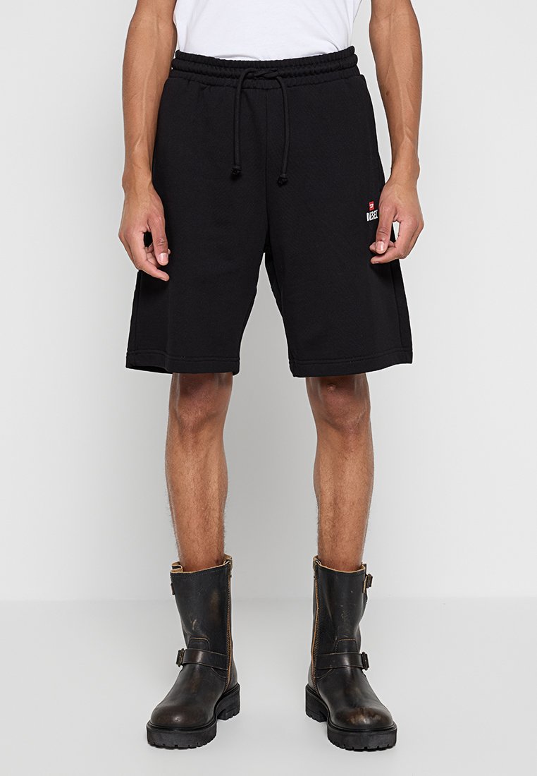 Mann trägt schwarze Shorts mit Kordelzug und schwarze Leder-Stiefeletten, steht vor einem einfarbigen Hintergrund.