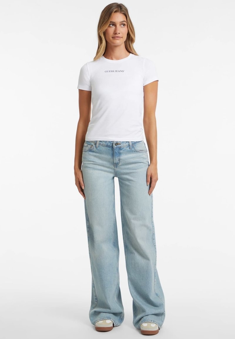 Weißes, tailliertes T-Shirt mit "GUESS JEANS"-Logo, kombiniert mit hellblauen, weit geschnittenen Jeans. Einfaches Design, Baumwollmaterial, minimalistischer Stil.