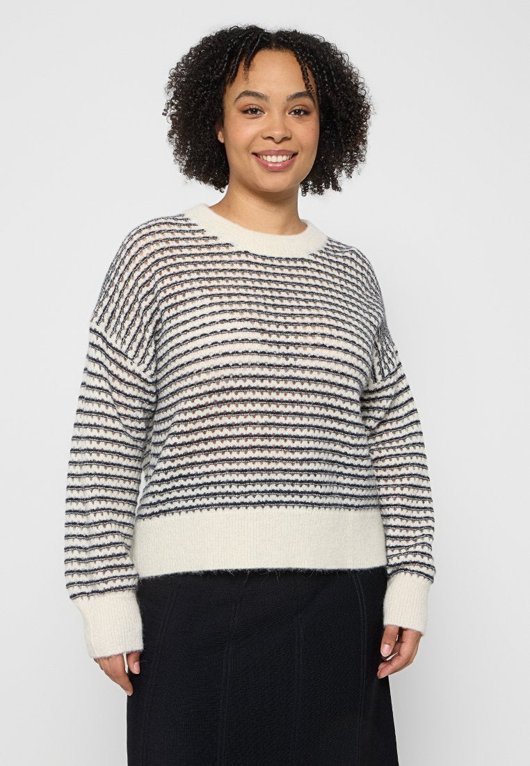 Frau mit lockigem Haar, die einen weißen Pullover mit schwarzen und braunen horizontalen Streifen und einen schwarzen Rock trägt, lächelt und vor einem grauen Hintergrund steht.