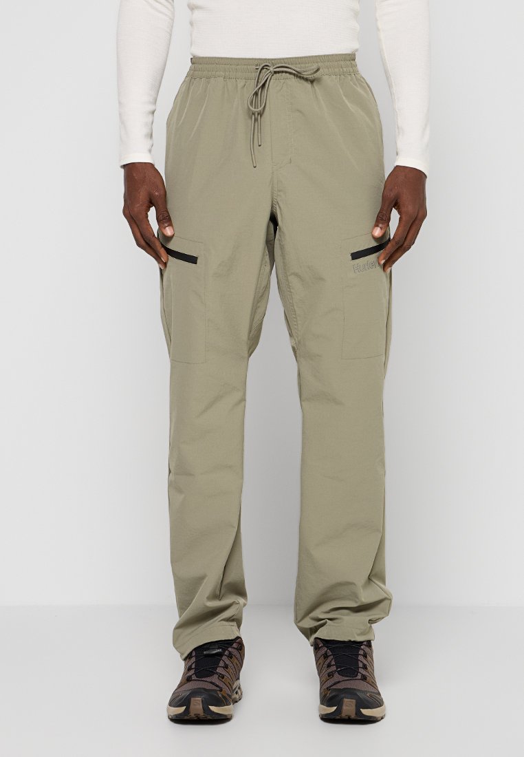 Helle khakifarbene Cargo-Hose mit elastischem Bund, Kordelzug und schwarzen Reißverschlusstaschen auf den Oberschenkeln. Getragen mit dunklen, gemusterten Schuhen.