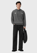 Grauer Hoodie mit Fronttasche, Logo-Akzenten und Kordelzügen, kombiniert mit schwarzen Jeans im weiten Schnitt und einer schwarzen Tasche.