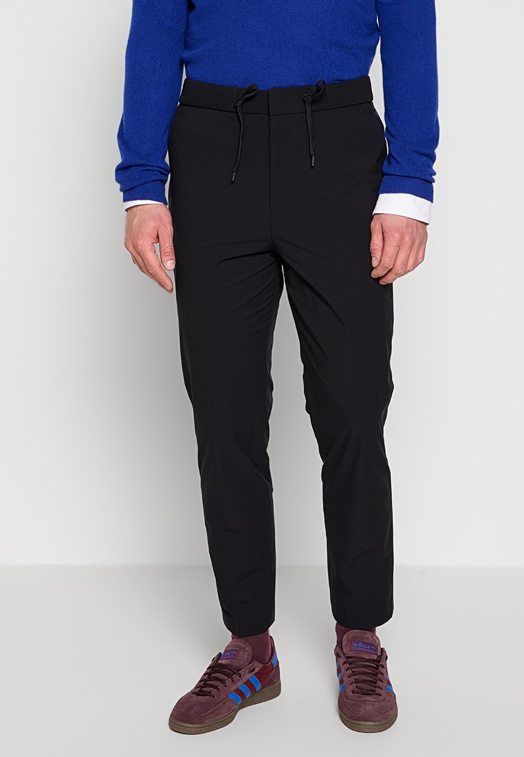 Schwarze tapered Jogginghosen mit elastischem Bund und Kordelzug. Kombiniert mit einem blauen Pullover und kastanienbraunen Sneakers mit blauen Streifen.