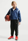 Junge Person in blauer Nike-Jacke, schwarzen Nike-Trainingshosen und leuchtend roten Nike-Schuhen, die einen Basketball hält und zur Seite schaut.