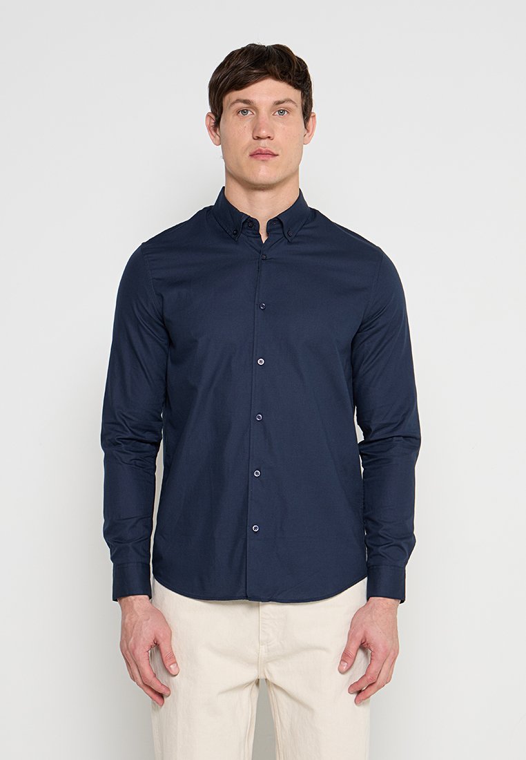 Mann trägt ein marineblaues Hemd mit langen Ärmeln und beige Hosen, steht vor einem schlichten hellen Hintergrund.