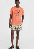 Korallenfarbenes T-Shirt mit schwarzem Grafikdruck, kombiniert mit cremefarbenen Shorts mit einem bunten tropischen Muster. Schwarze Flip-Flops runden das Outfit ab.