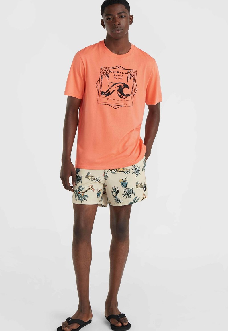 Korallenfarbenes T-Shirt mit schwarzem Grafikdruck, kombiniert mit cremefarbenen Shorts mit einem bunten tropischen Muster. Schwarze Flip-Flops runden das Outfit ab.