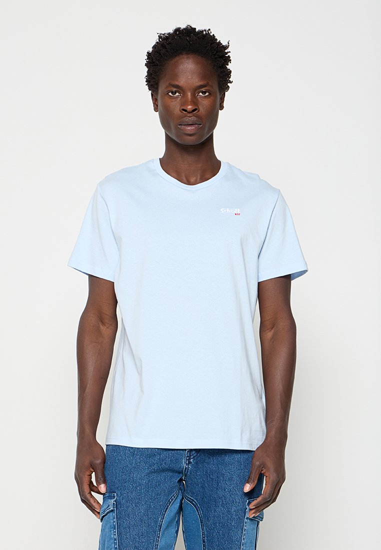 Mann mit kurzen lockigen Haaren, der ein hellblaues T-Shirt und blaue Jeans trägt, steht vor einem einfarbigen hellen Hintergrund.