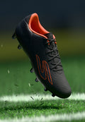 Schwarzer Fußballschuh mit strukturierter Oberfläche, orangefarbenen Akzenten und Schnürdesign, dargestellt vor einem Hintergrund aus grünem Gras mit fliegenden Rasenteilen.