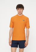 Junger Mann mit lockigem Haar, der ein orangefarbenes Kurzarmhemd und marineblaue Shorts trägt, steht vor einem schlichten weißen Hintergrund.