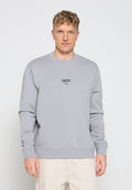 Mann trägt hellgrauen Pullover mit kleinem schwarzem Text auf der Brust und cremefarbenen Hosen, steht vor einem einfarbigen weißen Hintergrund.