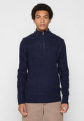 Junger Mann in einem strukturierten navyblauen Pullover mit einem kurzen Reißverschluss vorne und beigen Hosen, der vor einem schlichten, hellen Hintergrund steht.