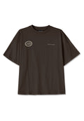 Dunkelbraunes, oversized T-Shirt aus Baumwolle mit einem runden Patch auf der linken Brust, der ein Hausdesign und das Wort "PEGADOR" in hellgrau zeigt.