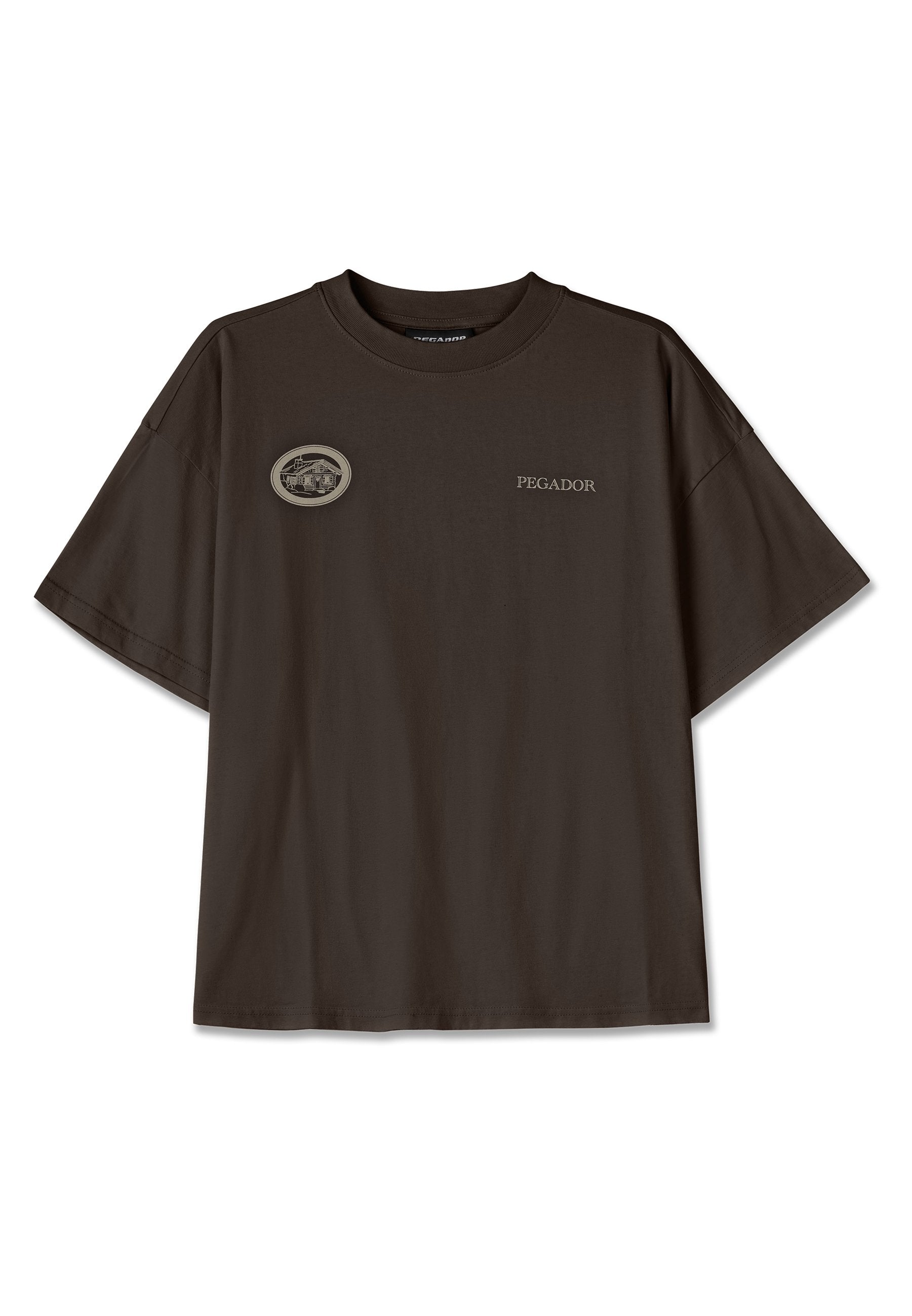 Dunkelbraunes, oversized T-Shirt aus Baumwolle mit einem runden Patch auf der linken Brust, der ein Hausdesign und das Wort "PEGADOR" in hellgrau zeigt.