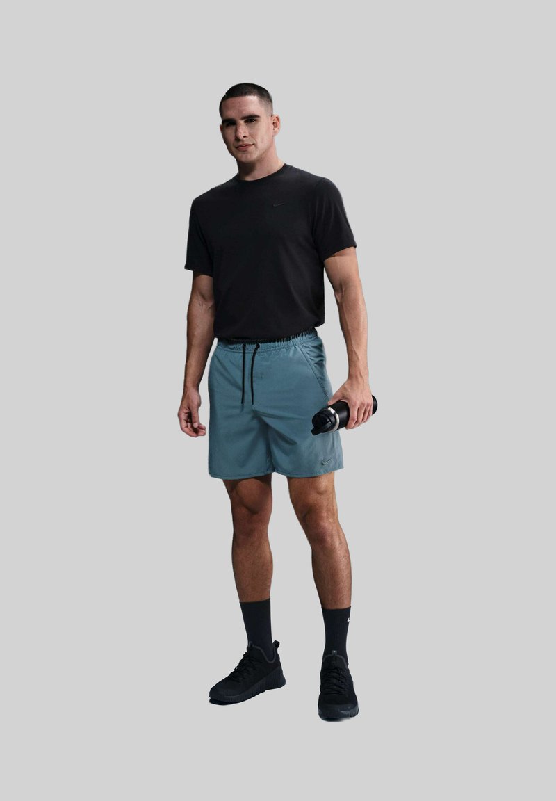 Schwarzes Kurzarmhemd für Männer, türkisfarbene Shorts mit elastischem Bund und Kordelzug, schwarze Wasserflasche und schwarze Sneakers. Glattes Material, sportliches Design.