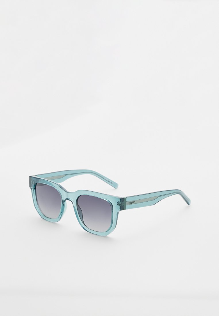 Quadratische blaue transparente Sonnenbrille mit dunklen Verlaufs-Gläsern, leicht schräg auf weißem Hintergrund gezeigt.