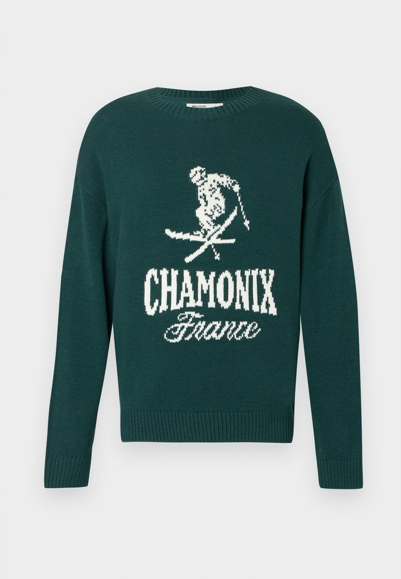 Grüner Strickpullover mit einer weißen Skifahrer-Grafik und dem Schriftzug "CHAMONIX Frankreich". Rippbündchen am Hals und Saum runden das Design ab.