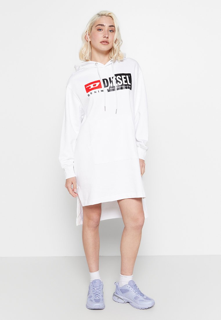Frau, die ein weißes Diesel-Hoodie-Kleid mit Fronttasche trägt, steht mit helllila Sneakers und weißen Socken vor einem schlichten Hintergrund.