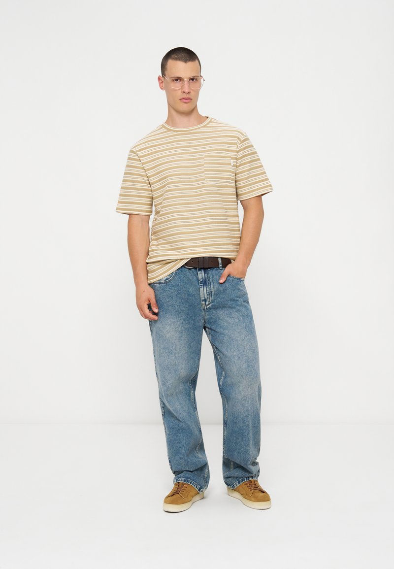 Mann mit kurzen Haaren und Brille, der ein beiges gestreiftes T-Shirt, blaue Jeans, einen braunen Gürtel und braune Turnschuhe trägt und vor weißem Hintergrund steht.