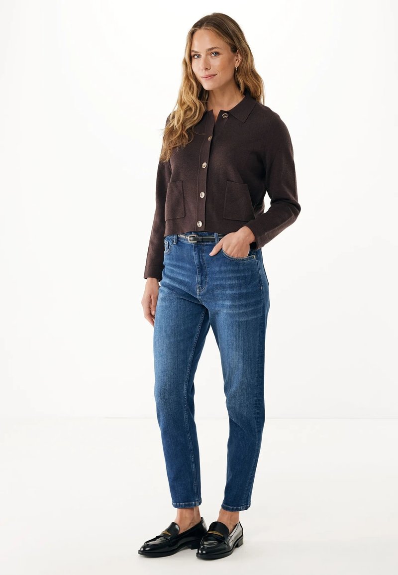 Frau mit langen, welligen Haaren, die eine dunkelbraune, geknöpfte Strickjacke, blaue Jeans, schwarzen Gürtel und schwarze Loafers trägt und mit einer Hand in der Tasche steht.