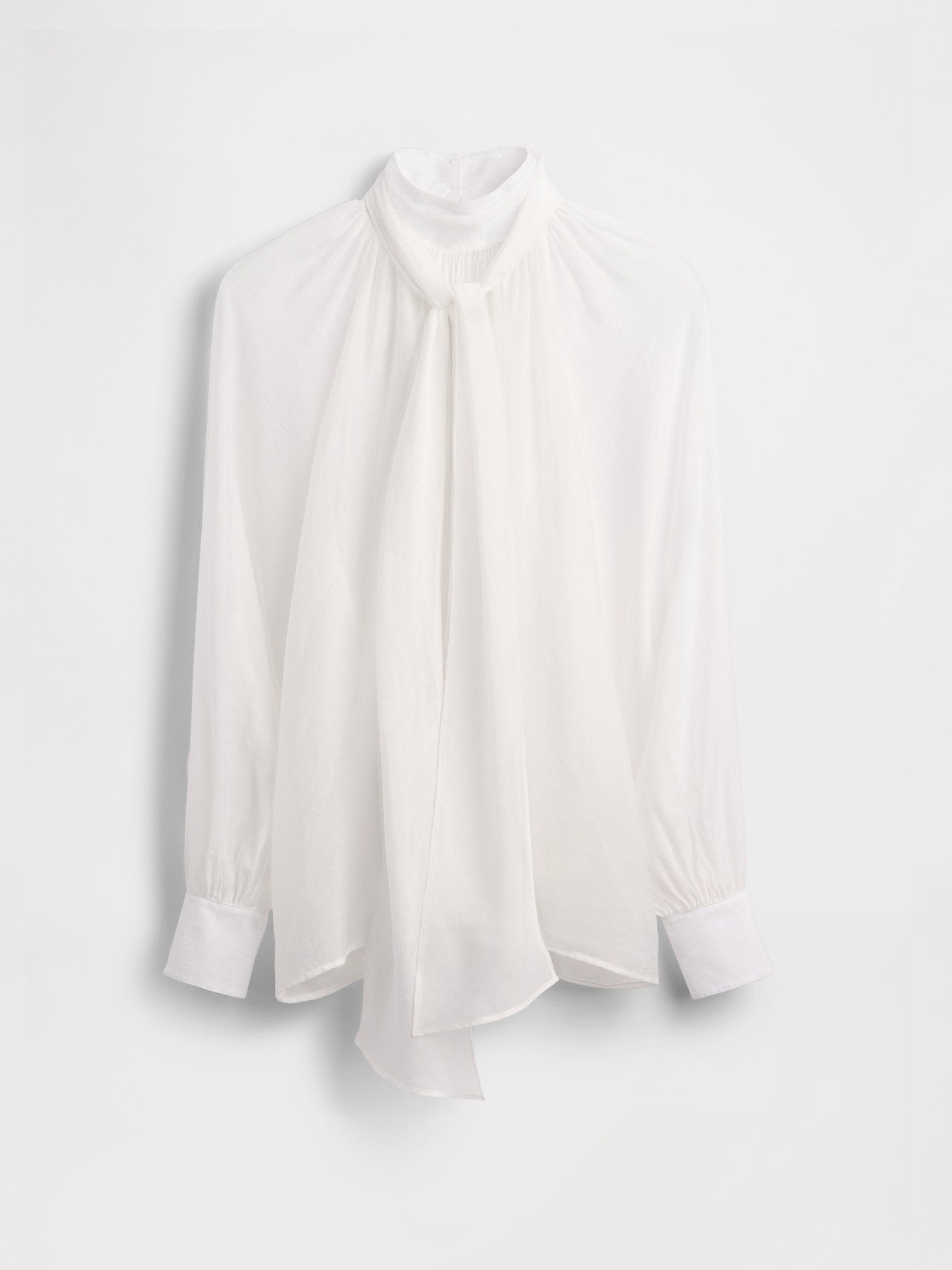 Weiße Bluse mit hohem Kragen, vorne fließend, lange Ärmel und geknöpfte Manschetten. Hergestellt aus einem leichten, transparenten Stoff.