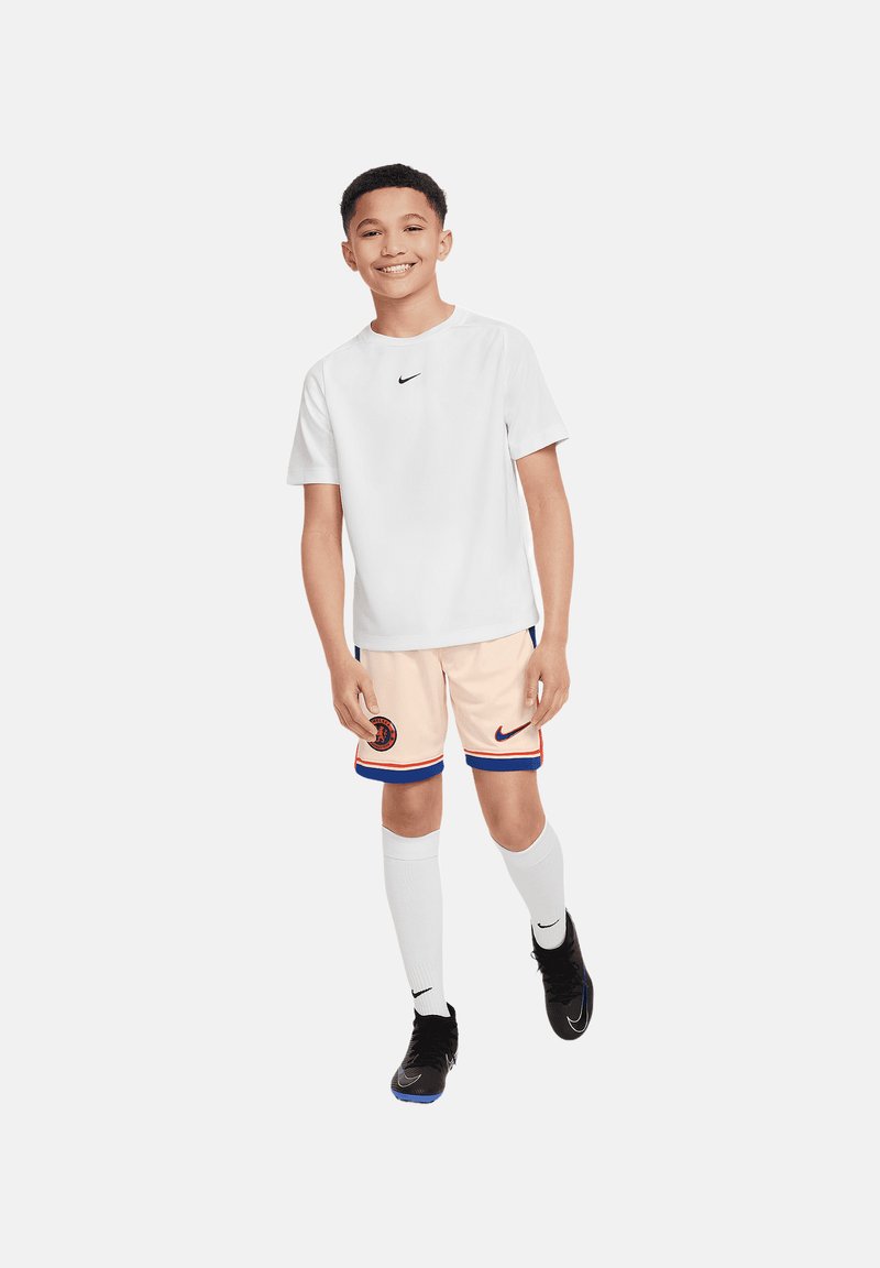 Weißes Sport-T-Shirt mit kleinem schwarzem Logo, pfirsichfarbene Shorts mit blauen Akzenten und Logo, weiße knielange Socken und schwarze Sportschuhe.