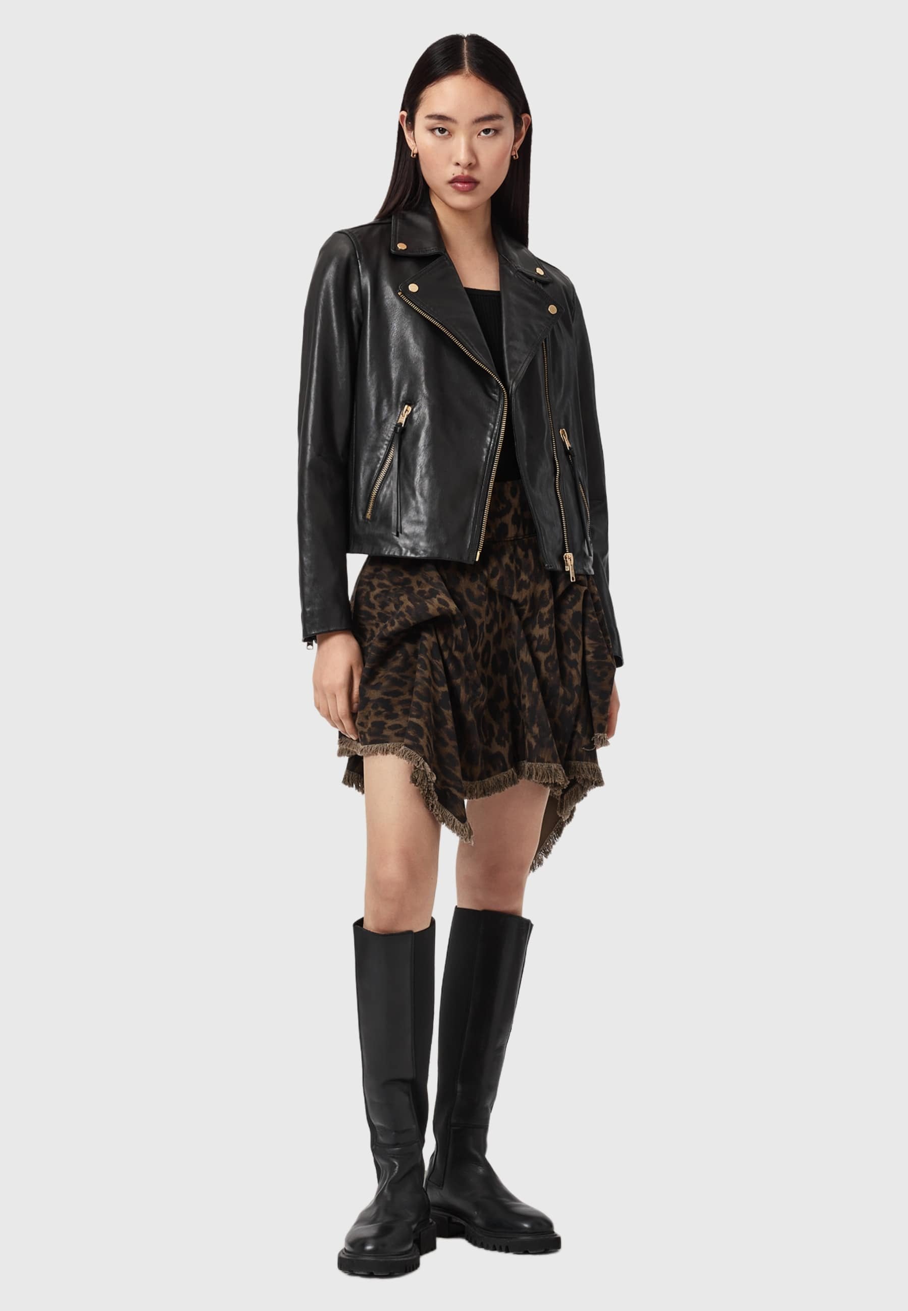 Schwarze Leder-Bikerjacke mit Reißverschlüssen, getragen über einem Leopardenmuster-Rüschrock. Ein Paar kniehohe schwarze Stiefel komplettiert das Outfit.