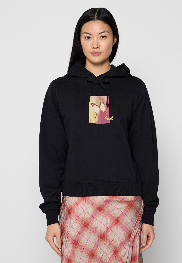 Eine Frau trägt einen schwarzen Hoodie mit einem Schmetterlingsmotiv auf der Brust und einen rot-weißen karierten Rock. Sie steht vor einem einfarbigen hellen Hintergrund.