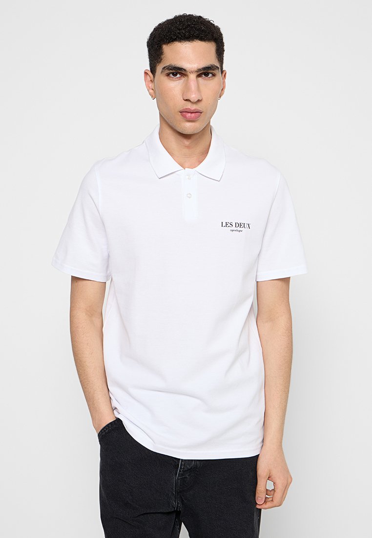 Weißes Polo-Shirt mit kurzen Ärmeln und einer Knopfleiste mit drei Knöpfen. Es verfügt über ein kleines schwarzes Logo auf der linken Brust und eine glatte Baumwolloberfläche.
