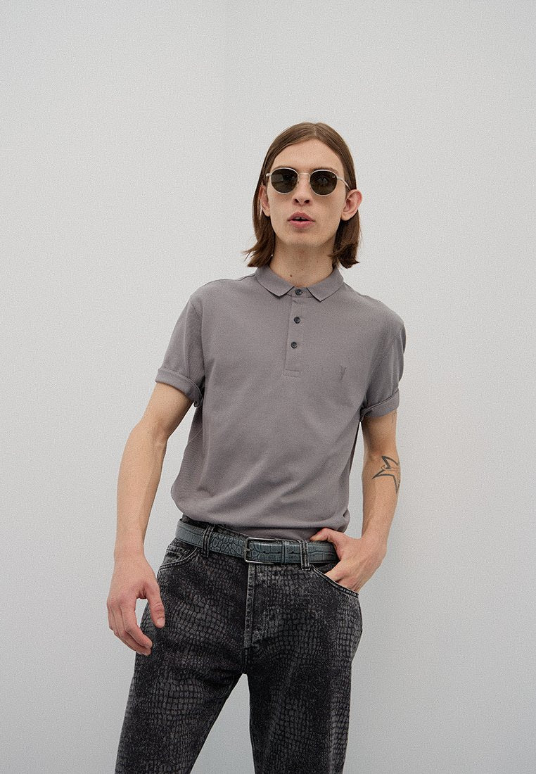Graues Polohemd mit Kragen und drei Knöpfen, kombiniert mit strukturierten schwarzen Jeans. Die Accessoires umfassen runde Sonnenbrillen und einen gemusterten Gürtel.