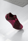 Dunkelmaroonfarbener Sportschuh mit rosa Akzenten. Merkmale sind ein Mesh-Obermaterial, eine abgerundete Zehenpartie und eine strukturierte Sohle. Schnürsenkel und Logo deutlich sichtbar.