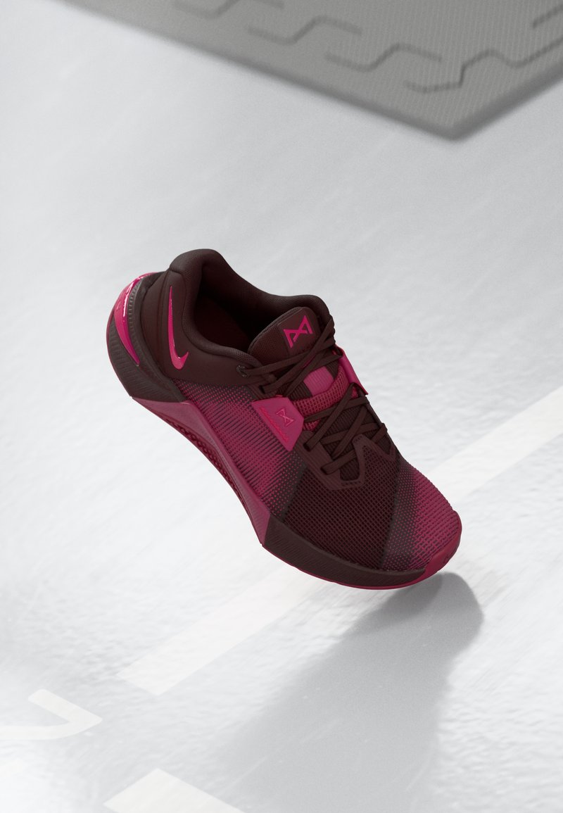Dunkelmaroonfarbener Sportschuh mit rosa Akzenten. Merkmale sind ein Mesh-Obermaterial, eine abgerundete Zehenpartie und eine strukturierte Sohle. Schnürsenkel und Logo deutlich sichtbar.