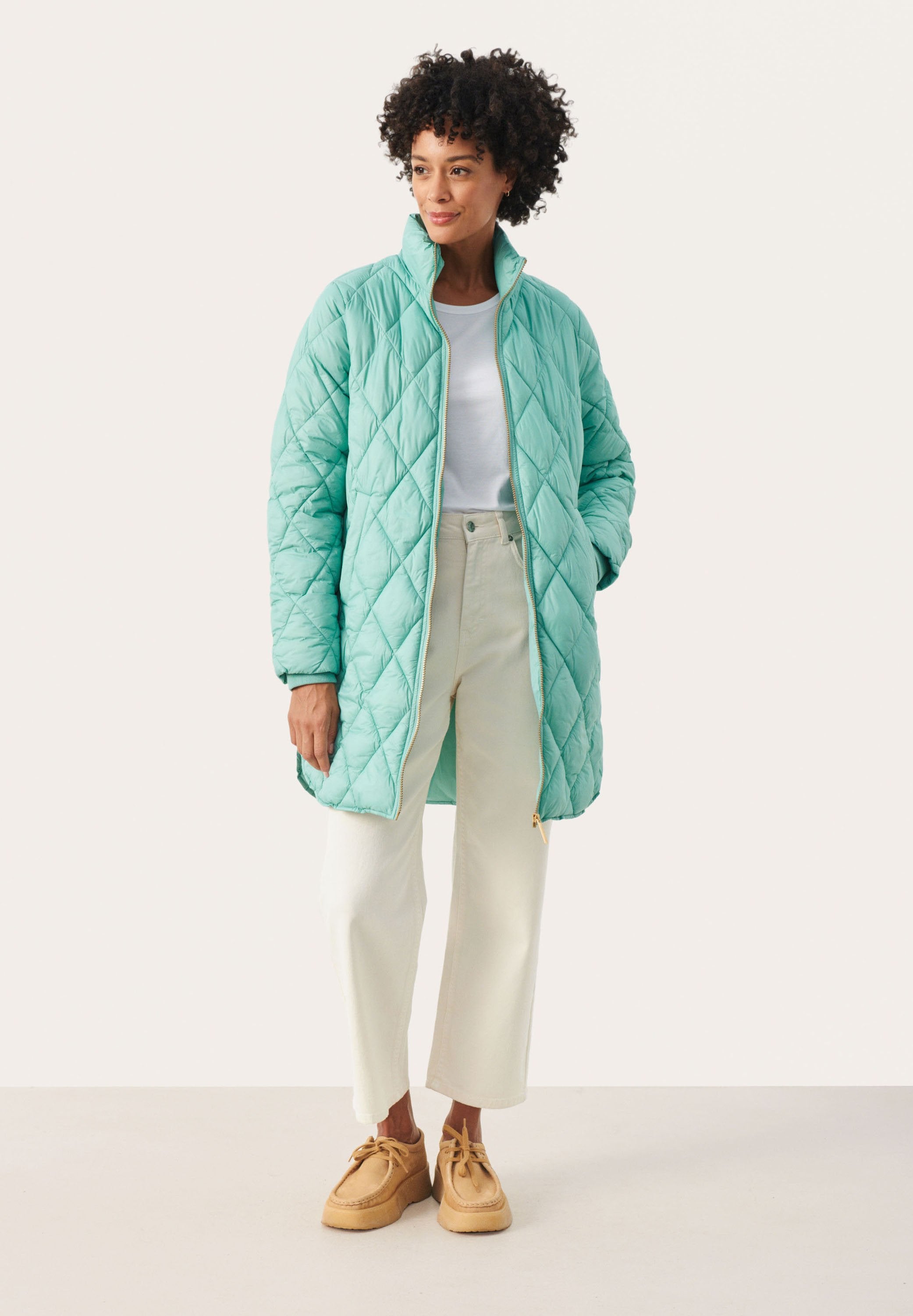 Steppjacke in Türkis mit hohem Kragen, goldfarbenem Reißverschluss und Seitentaschen, kombiniert mit hellbeigen Hosen und braunen Mokassins.