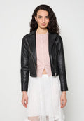 Frau mit lockigem Haar, die eine schwarze Lederjacke über einem rosa, geknöpften Oberteil und einem weißen Spitzenrock trägt, steht vor einem schlichten hellen Hintergrund.